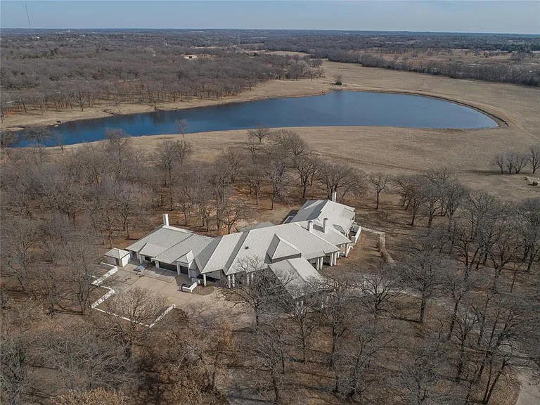 Aerial view of a home surrounded by trees, open land, and a pond